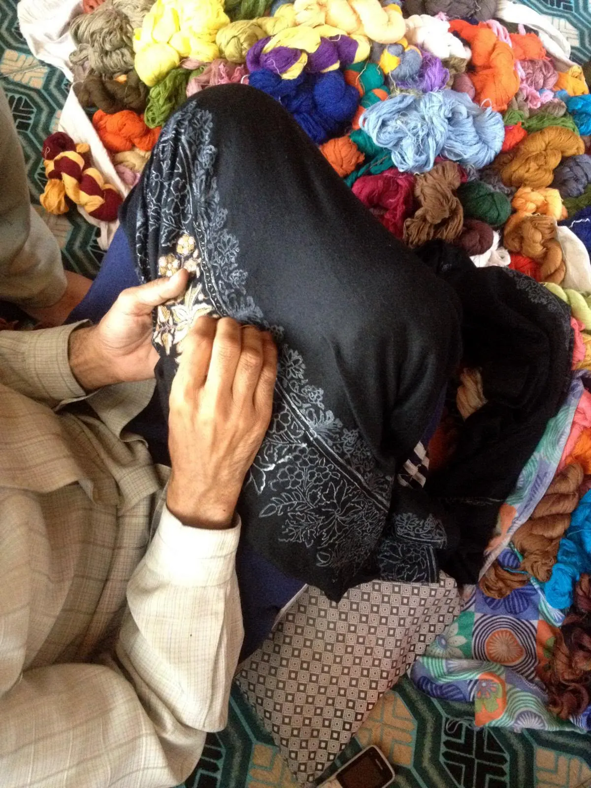 An embroiderer working ivory needlework into a black shawl, surrounded by coloured yarn.