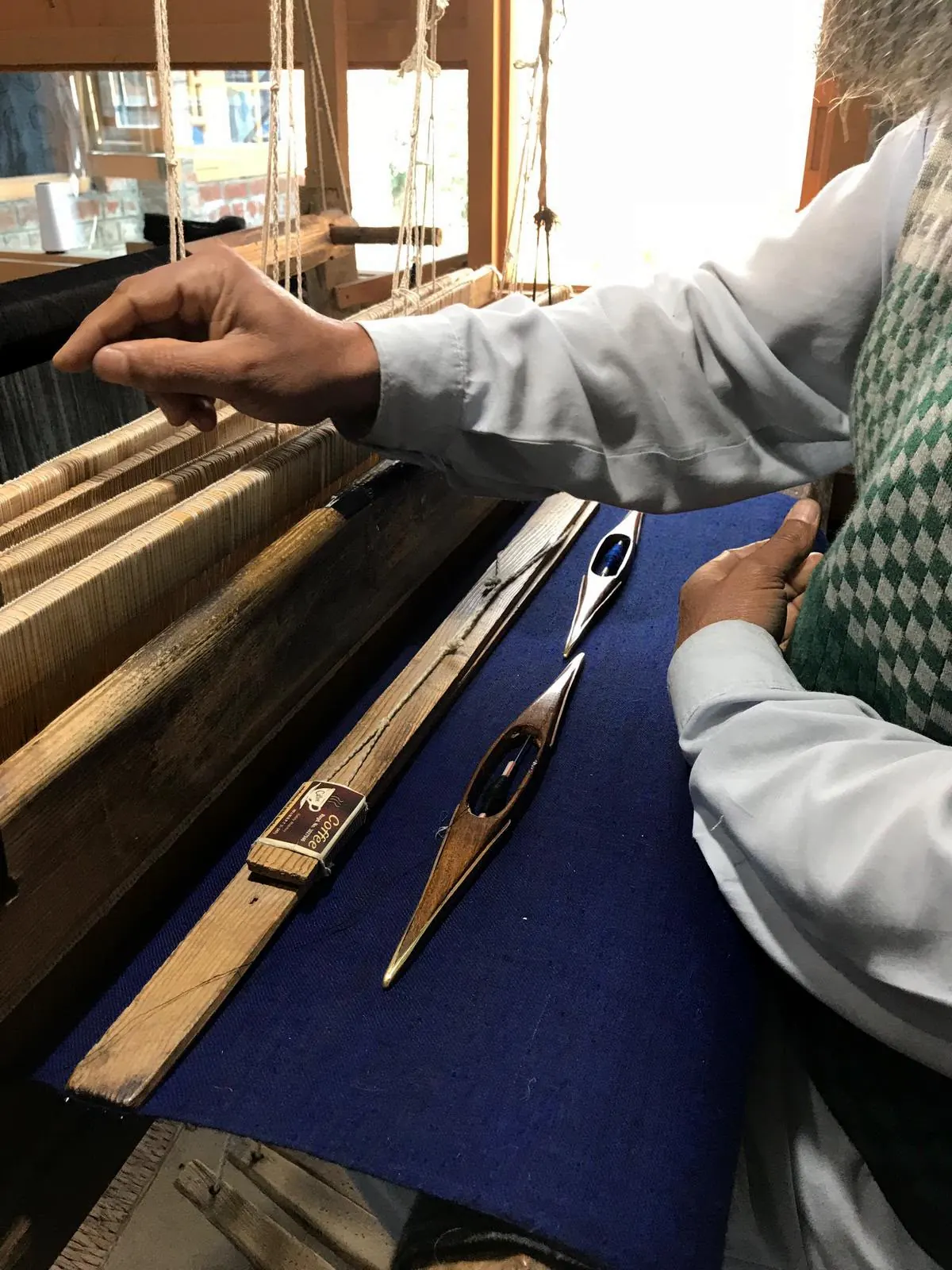 A weaver's hands on a wooden loom, two shuttles lying on the dark blue cloth.
