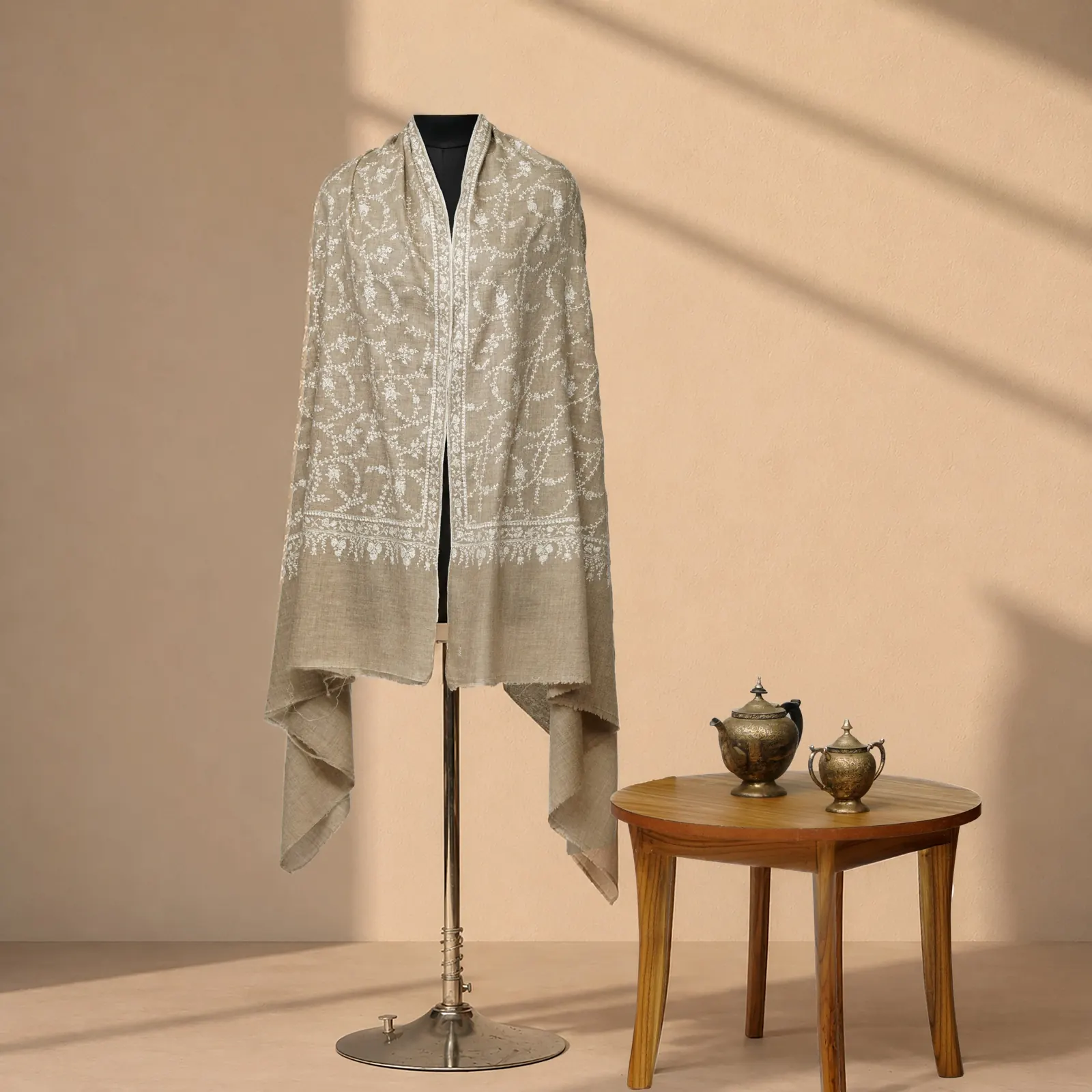 Oatmeal shawl with cream floral needlework running down a centre panel, beside brass teapots.