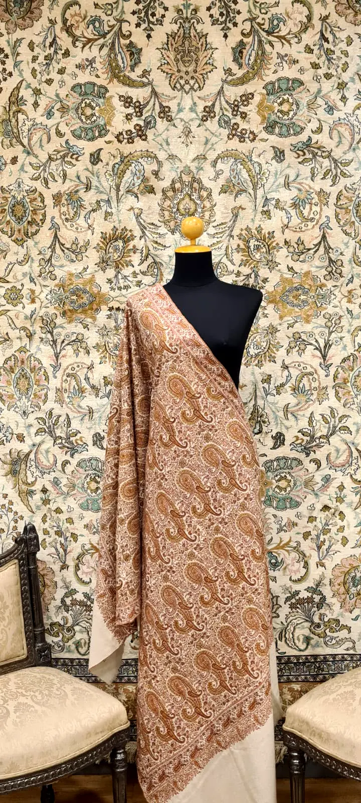 Shawl covered in dense rose and peach needlework, draped on a stand before a patterned wall.