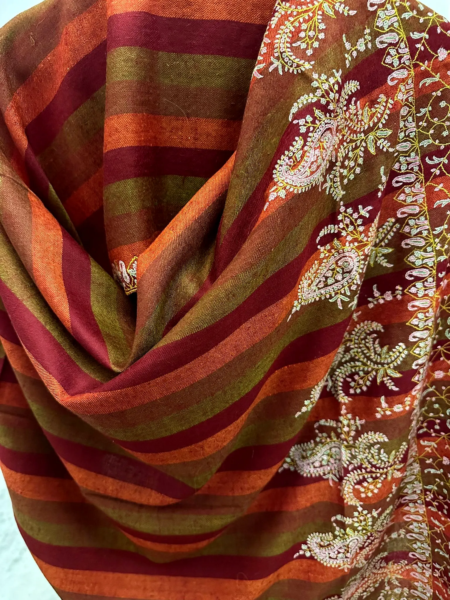 Shawl in red, orange and olive stripes, draped, with ivory paisley needlework along one edge.