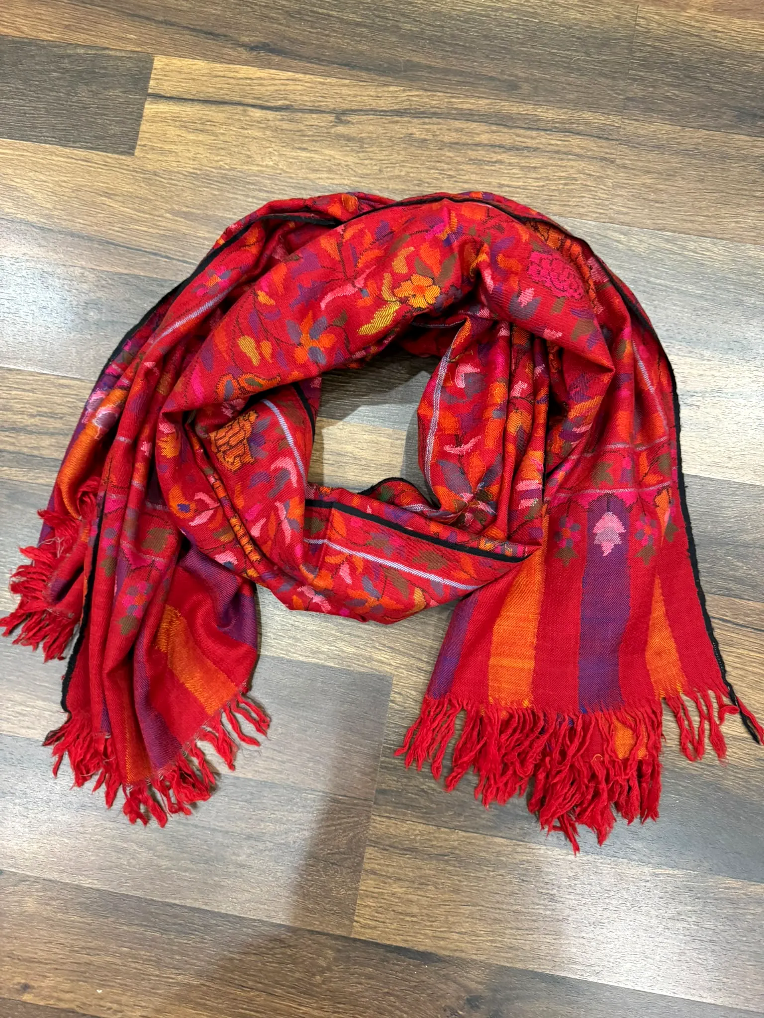 Scarlet kani stole with orange and pink motifs and a red fringe, laid on a wooden floor.